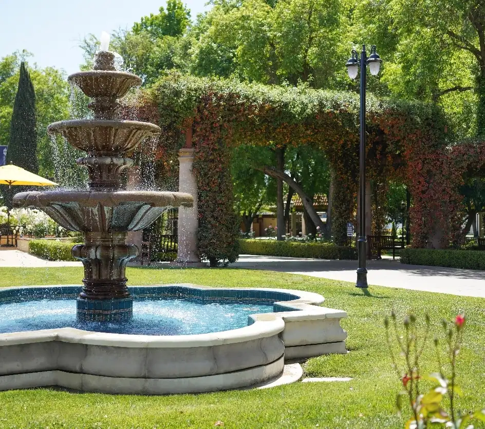 Photo of the quatrefoil fountain on the grounds. In the background are a yellow patio umbrella, an arbor covered in flowering vines, and an elegant streetlamp.